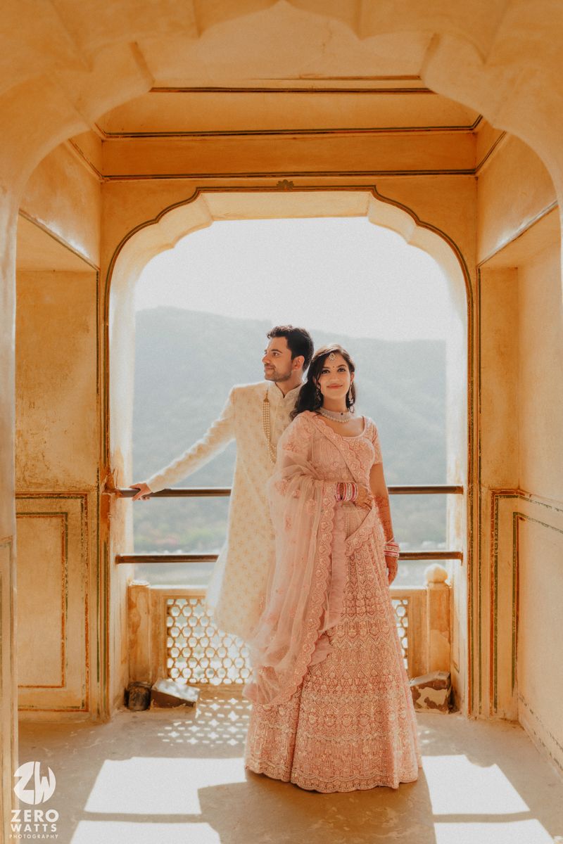 A couple poses in traditional Indian wedding attire against an ornate archway overlooking misty mountains.