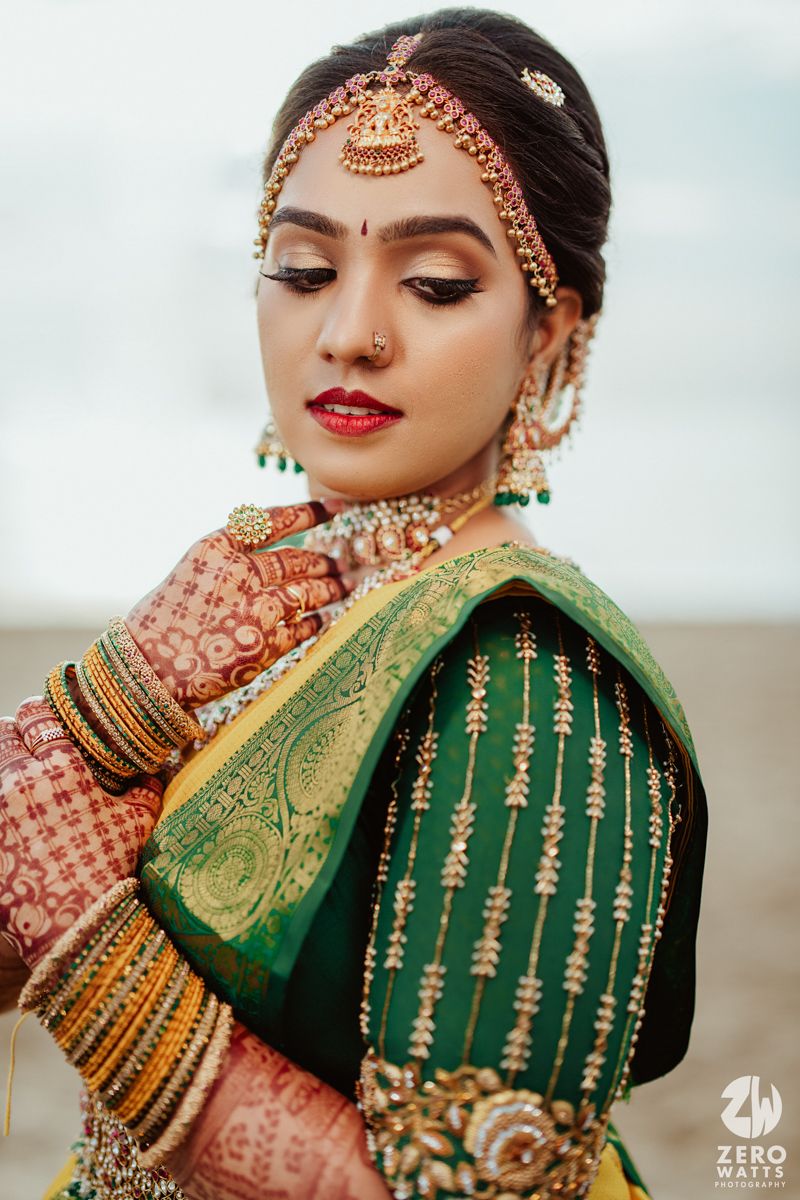 Close-up portrait of bride in green silk saree with traditional Indian bridal jewelry and makeup against beach backdrop.