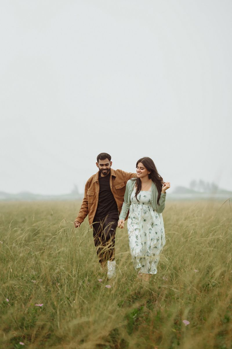 A couple walking together and laughing through a misty meadow of tall grass on an overcast day.