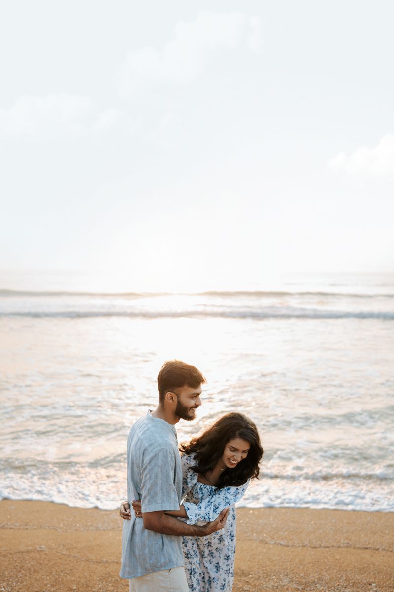 A romantic couple embraces on a sandy beach at sunset wearing coordinating blue and white outfits.
