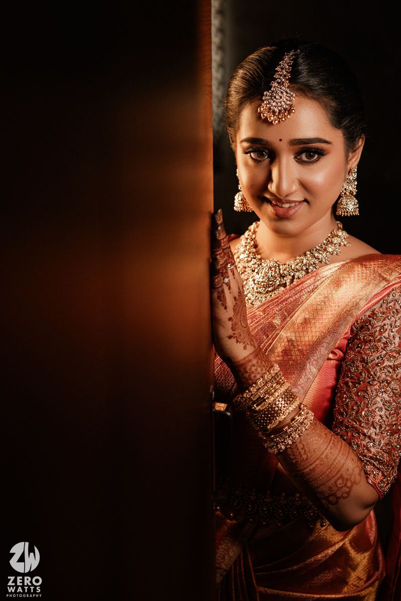 A bride in traditional South Indian wedding attire with ornate gold jewelry and a silk saree poses against a dark backdrop.