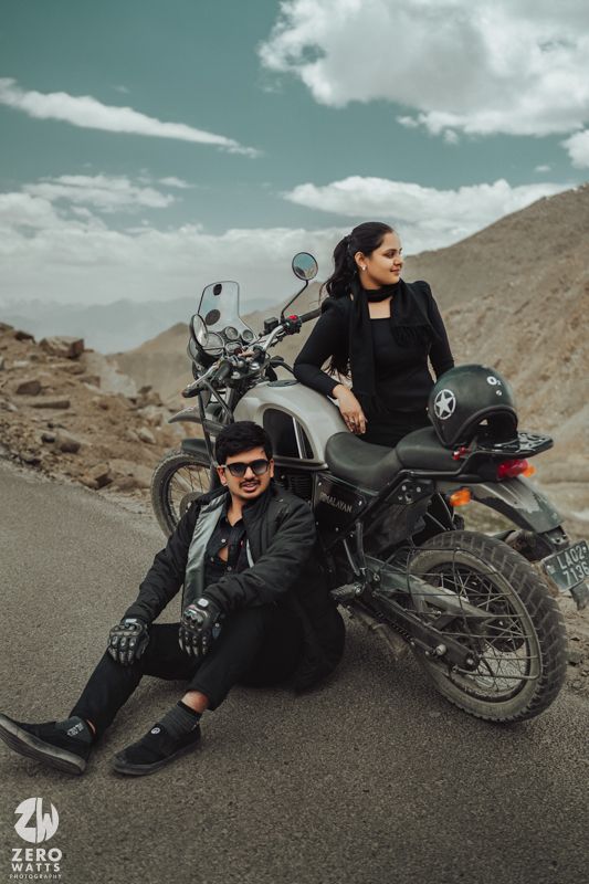 Black motorcycle parked on road with couple posing in dramatic mountain scenery.