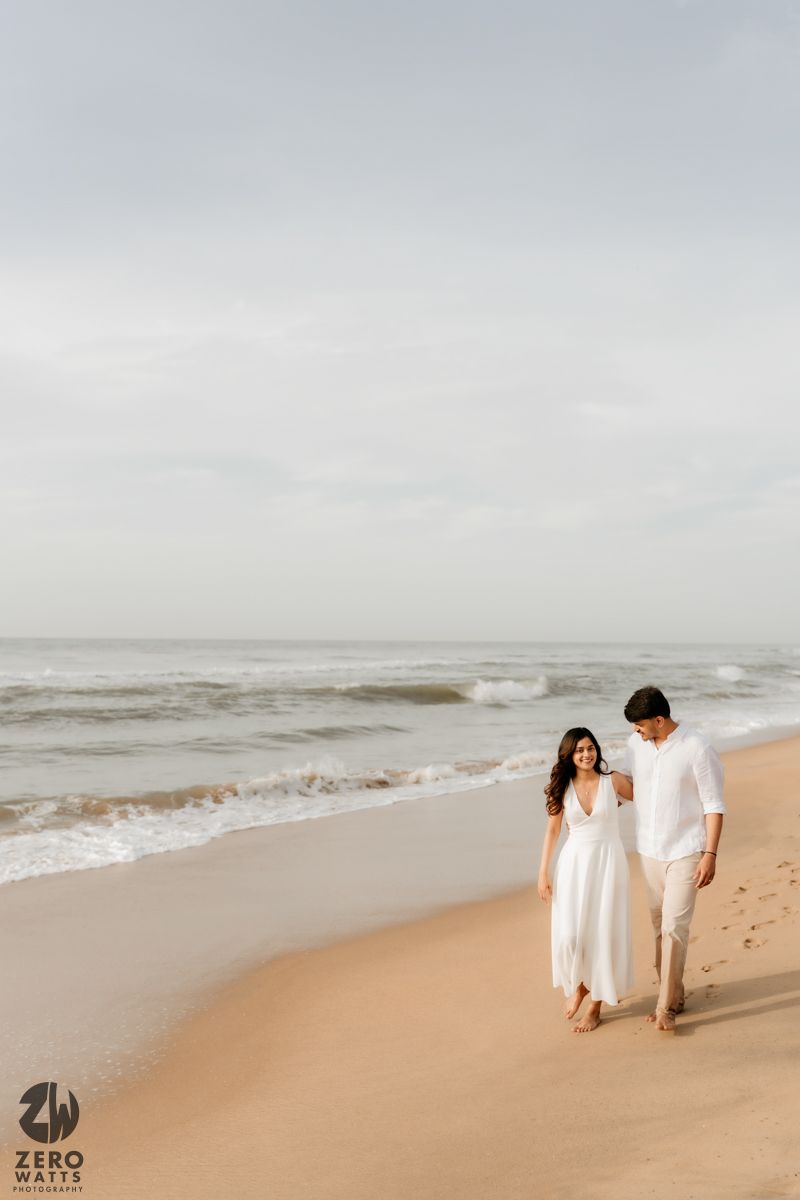 Romantic beach photoshoot with couple in white clothing walking along sandy shoreline.