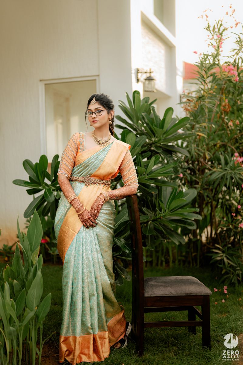 Someone wearing a mint green and peach silk saree poses elegantly next to a wooden chair in a garden setting.