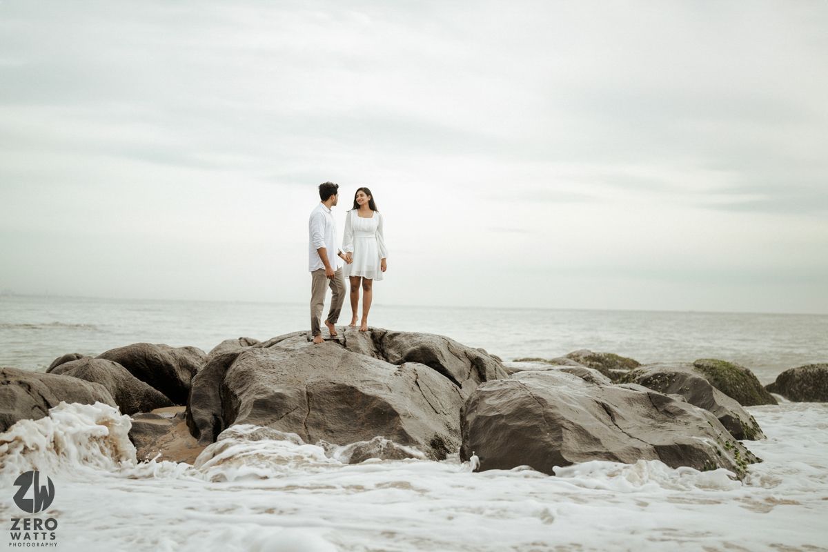 Two figures in white stand atop large rocks by the ocean shore as waves crash against the rocky coastline.