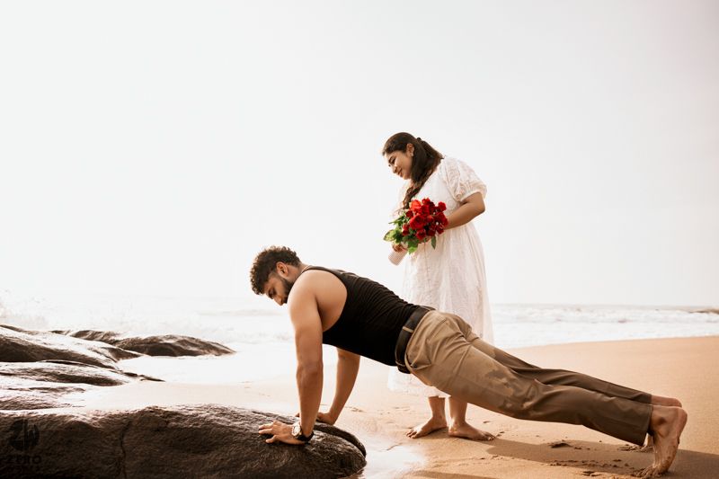 Couple on beach rocks with one person exercising while other holds flowers.