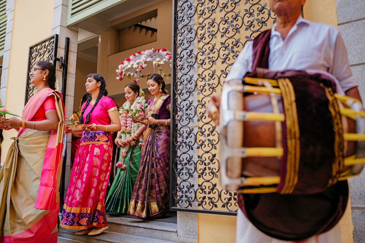 Colorful traditional Indian wedding celebration with musicians and guests in sarees standing outside a decorated venue.