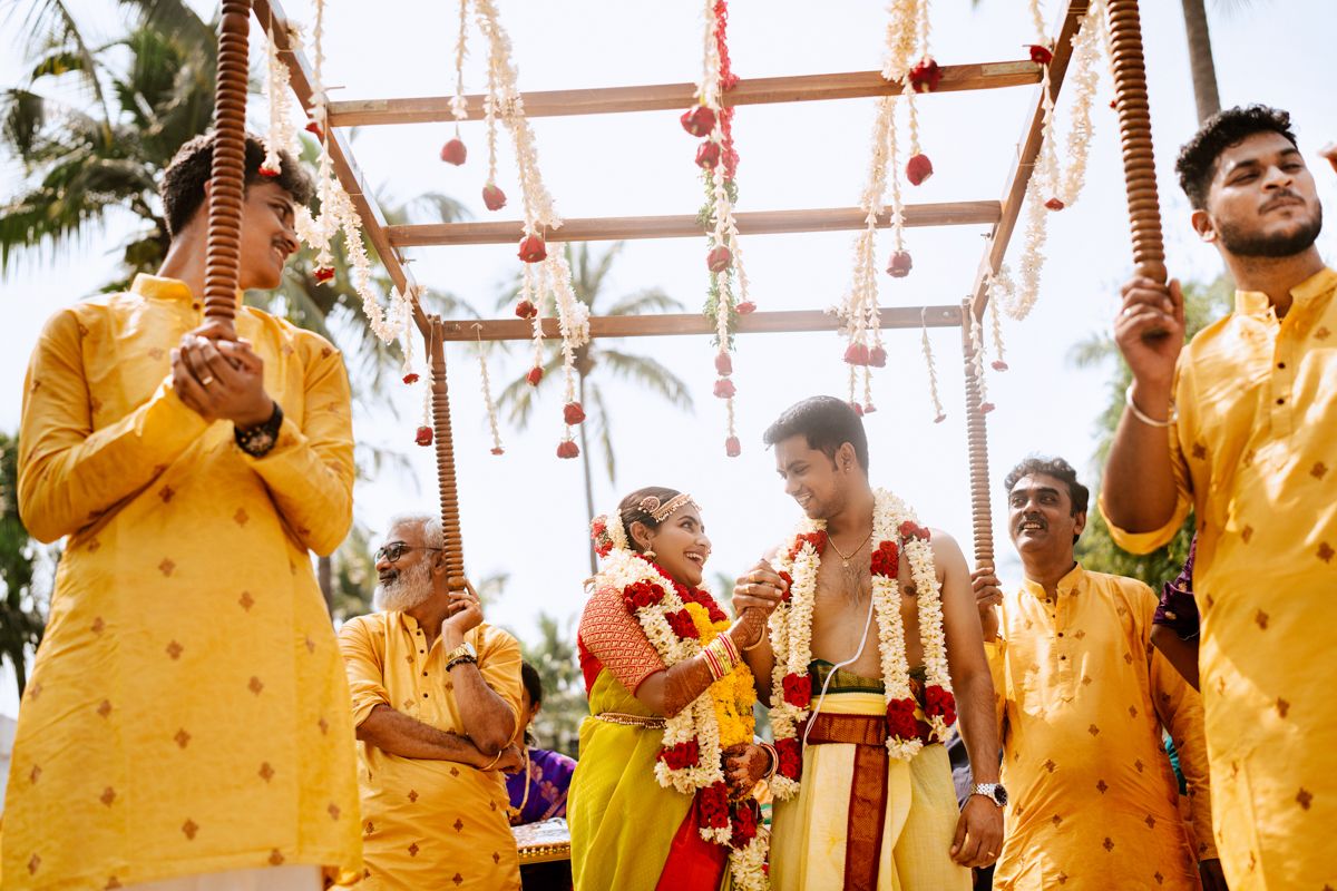 Group wearing yellow traditional outfits performing wedding rituals under decorated wooden mandap with palm trees.