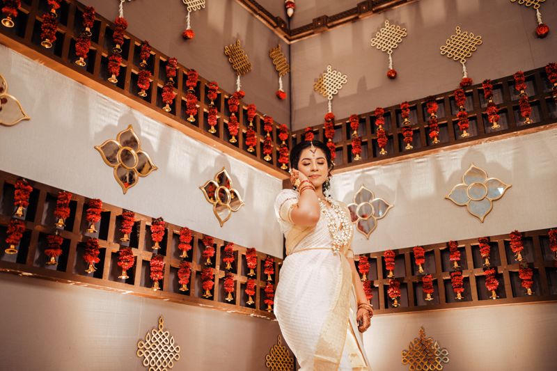 Traditional South Asian bridal scene with white sari against ornate backdrop featuring hanging lamps and geometric wall decor.
