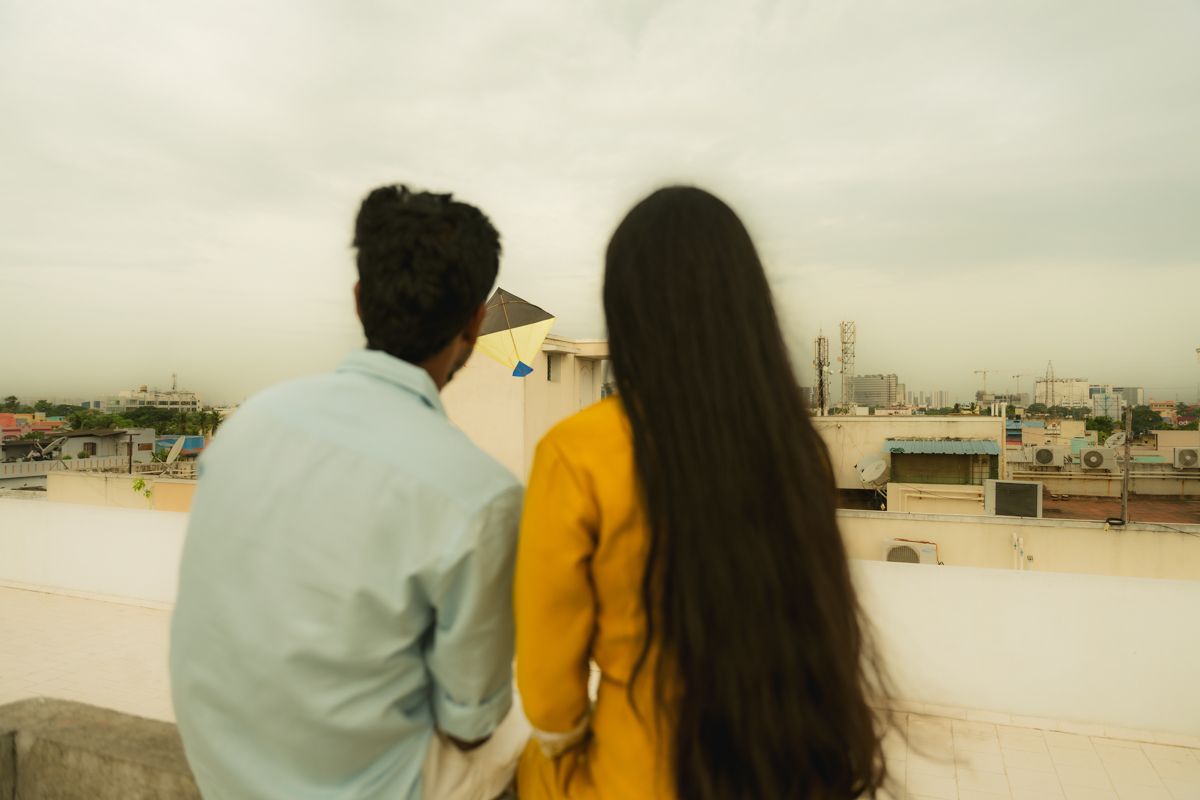 Two people in white and yellow outfits sit together overlooking a cityscape from a rooftop viewpoint.