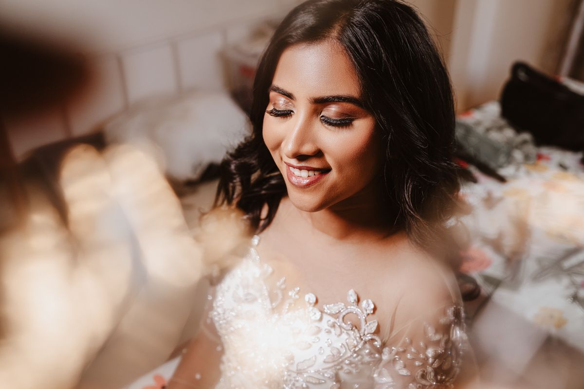 A smiling bride wearing a delicate lace wedding gown with floral embellishments looks down with closed eyes.