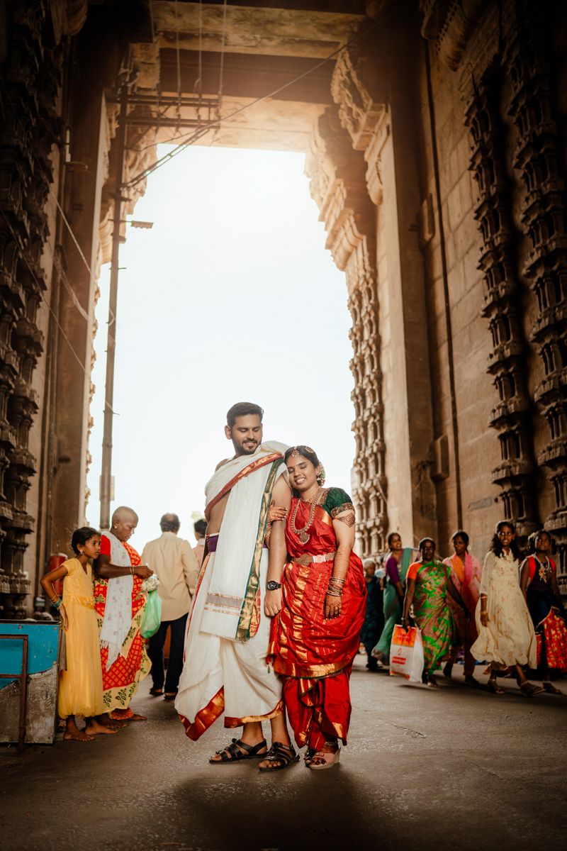 A couple in traditional Indian dress poses beneath an ornate archway while visitors walk through a temple corridor.