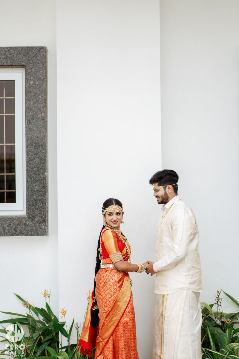 A couple in traditional Indian wedding attire pose together against a white wall with green plants.