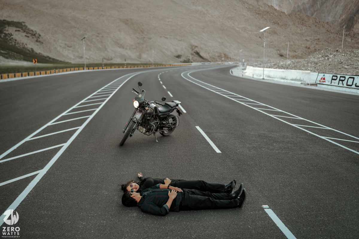 Two people lie on empty mountain road next to parked motorcycle creating artistic travel photography composition.