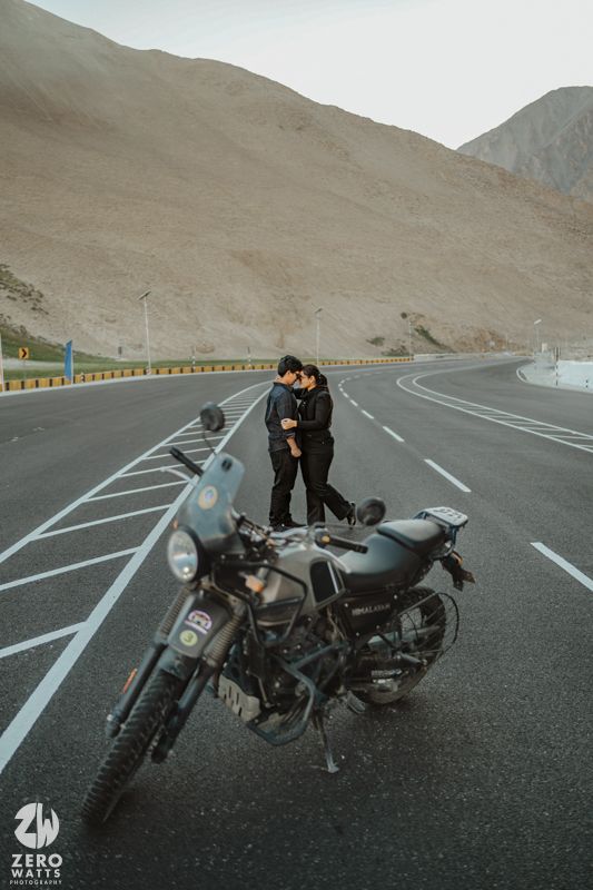 A parked motorcycle on a curving desert highway with two figures embracing near barren mountain slopes.