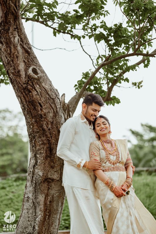 Romantic couple in traditional Kerala wedding attire embrace beneath a large tree in an outdoor setting.