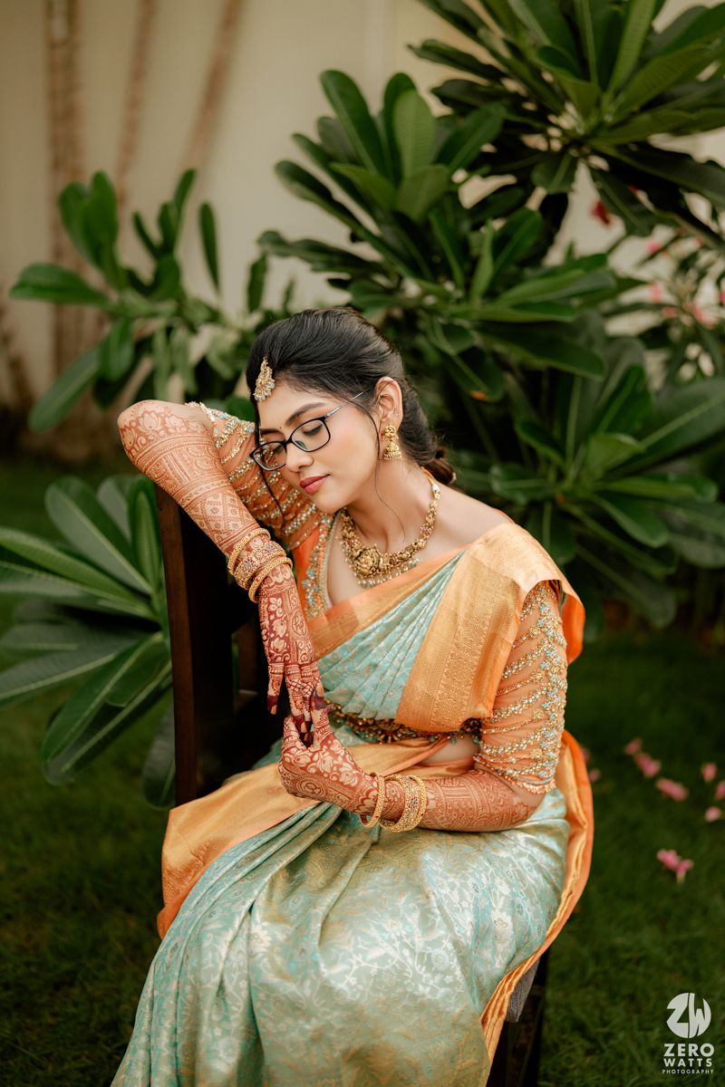 A person wearing a mint green and orange silk saree with intricate mehndi designs poses elegantly against tropical foliage.