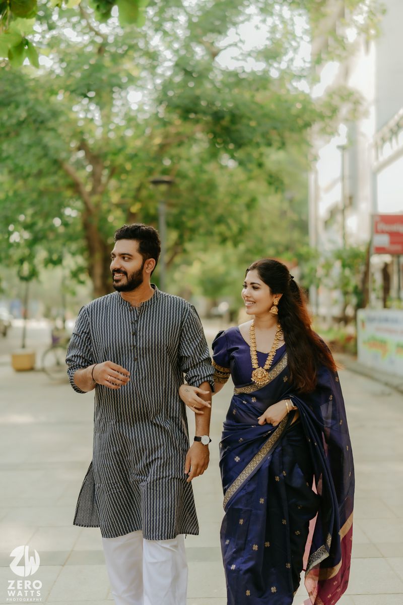 A couple walking and laughing together outdoors on a tree-lined path wearing traditional Indian attire.