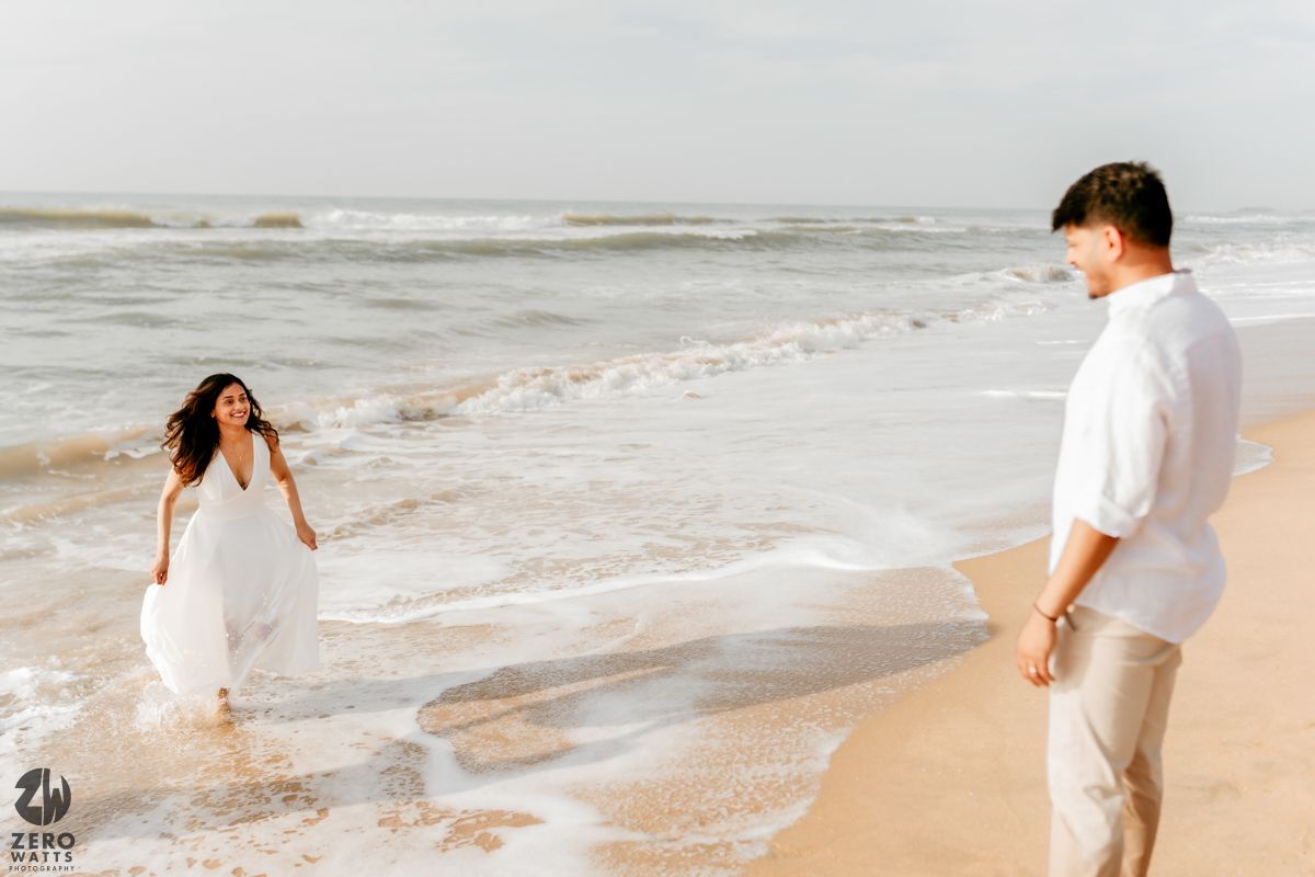 Couple in white clothing exploring beach as waves crash on shoreline during cloudy day.