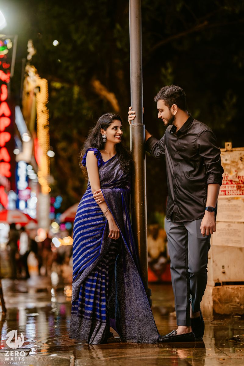 A stylish couple posing near a lamppost on a rainy evening with colorful lights in the background.