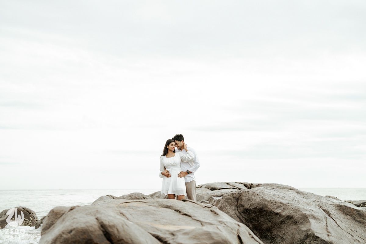 A romantic couple in white outfits embraces while standing on dramatic rocky coastal formations under an overcast sky.