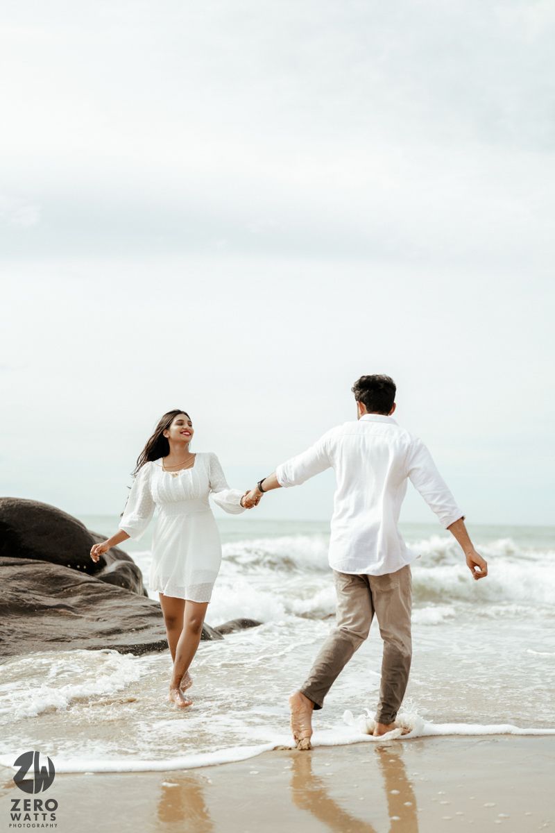 A couple in white clothing holds hands while running on a beach with crashing waves.
