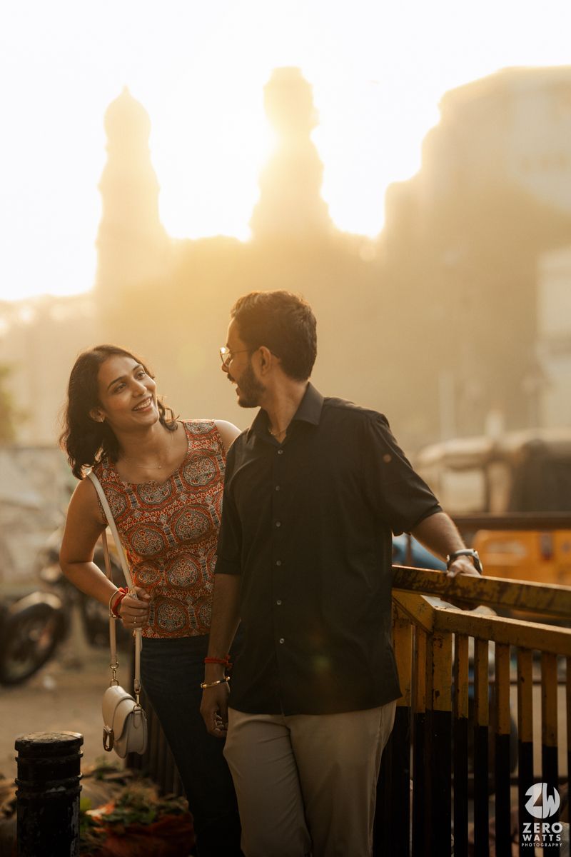 A couple shares a tender moment on a balcony during golden hour with city silhouettes behind them.
