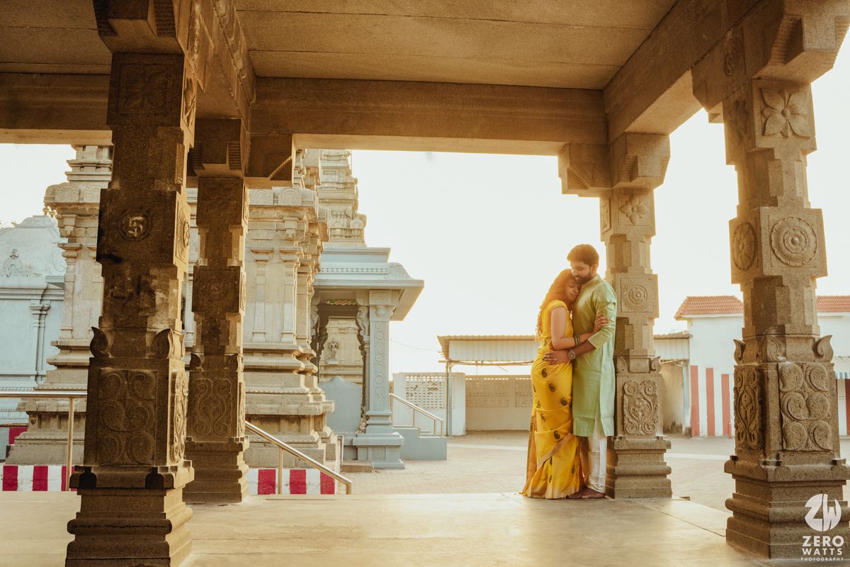 Couple in traditional Indian attire embracing under ornate temple pillars during golden hour.