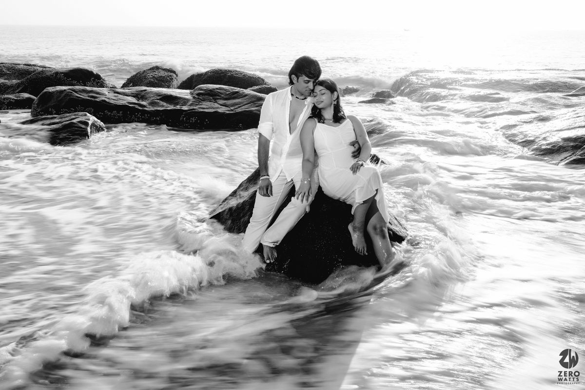 Black and white photo of couple sitting on seaside rock with waves surrounding them.