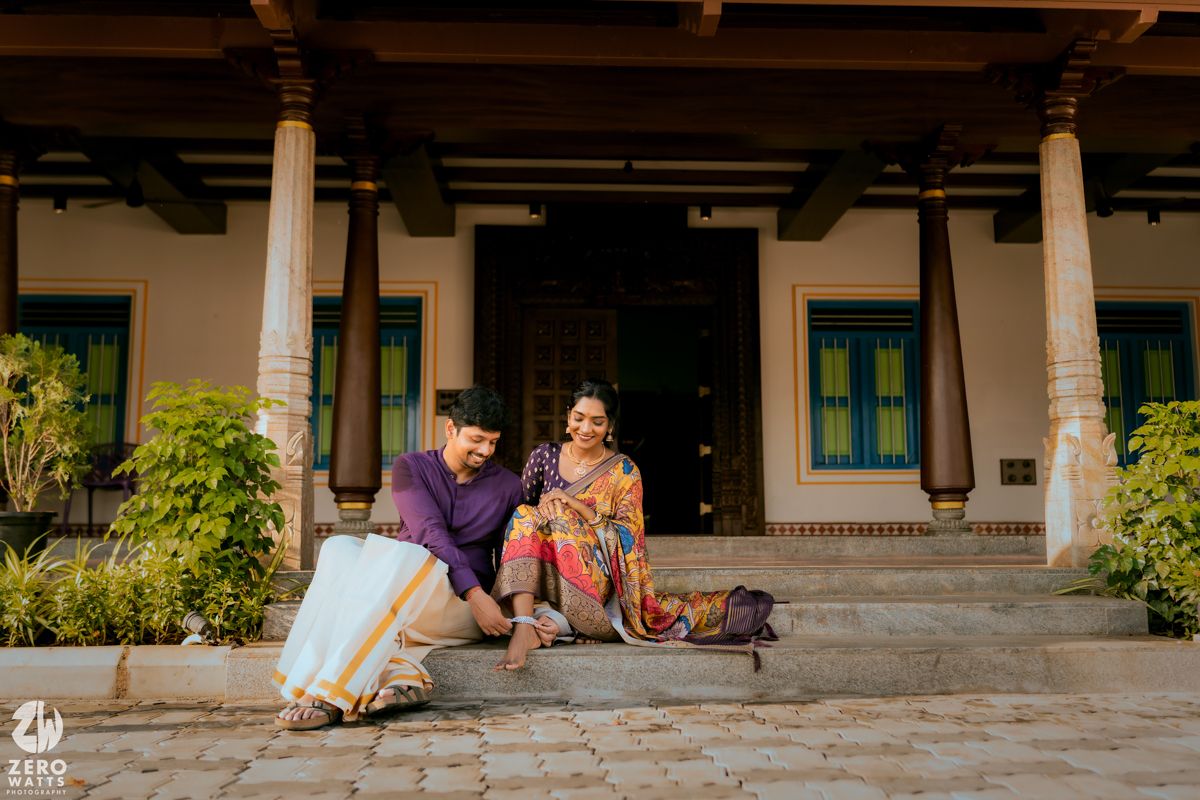 Two people in traditional attire sit relaxed on steps of a traditional South Indian home with white columns and greenery.