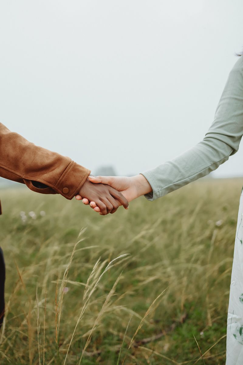 Two people hold hands tenderly in a field of tall grass against a misty background.