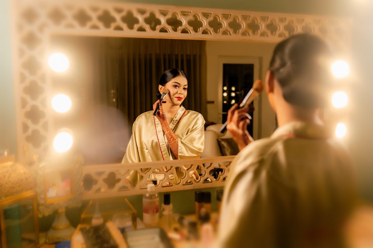 A person applies makeup while sitting at an illuminated vanity mirror in warm, glowing lighting.