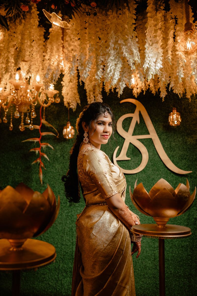 Traditional Indian engagement decor featuring hanging wisteria flowers, ornate gold monogram, and lotus-shaped holders in warm lighting.