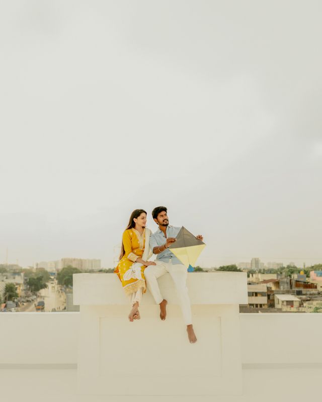 A couple sits casually on the edge of a white rooftop overlooking a cityscape in the distance.