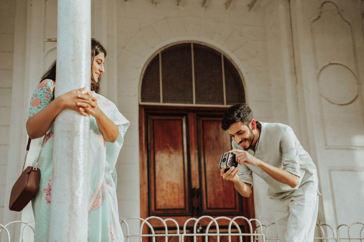 Two people in retro clothing pose playfully against white columns of a vintage building exterior.
