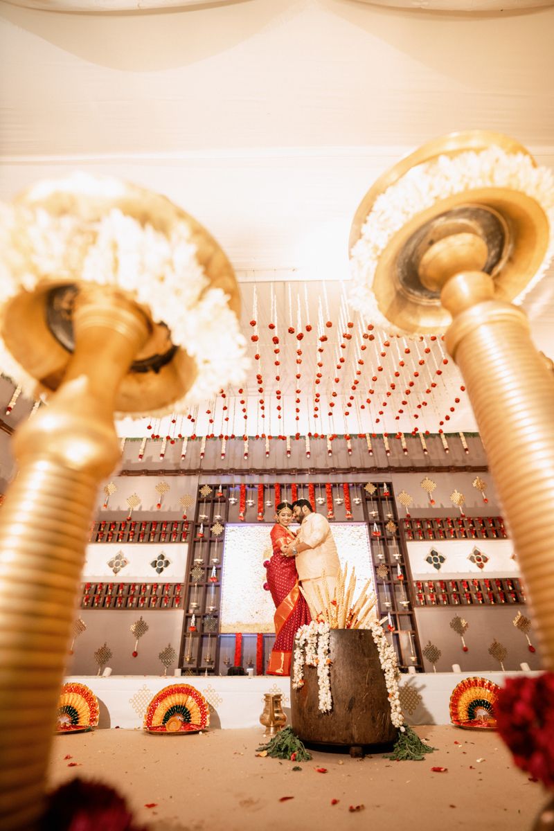 Artistic wedding photograph captured through decorative brass temple lamps with flower garlands.