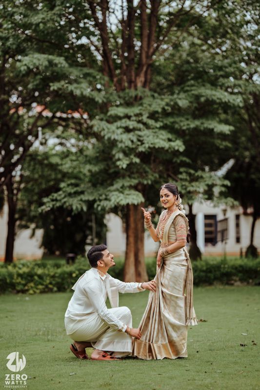 A couple in traditional South Indian attire shares a romantic moment on a lush green lawn surrounded by trees.