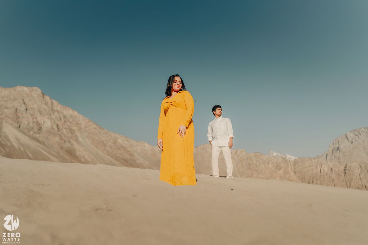 A couple in yellow dress and white attire pose against mountainous desert landscape at sunset.