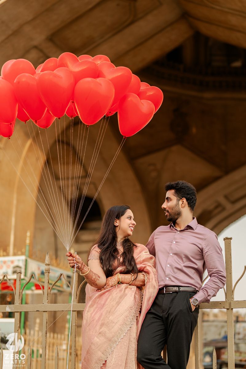 A couple poses with red heart-shaped balloons under an elegant architectural structure.