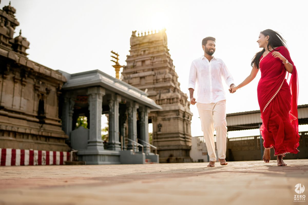 Two people share a joyful moment while walking together outside a traditional Indian temple with ornate architecture in the background.
