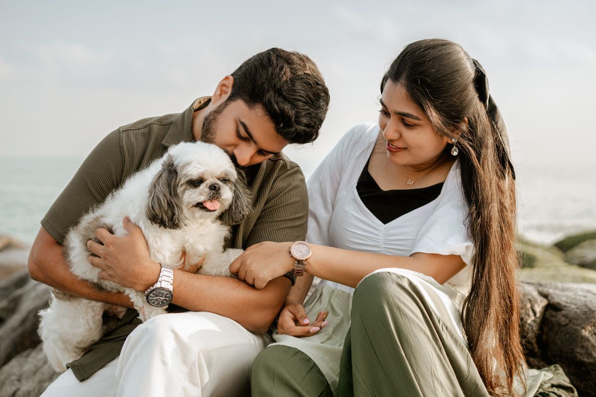 A couple sitting on coastal rocks while petting their small white Shih Tzu dog.