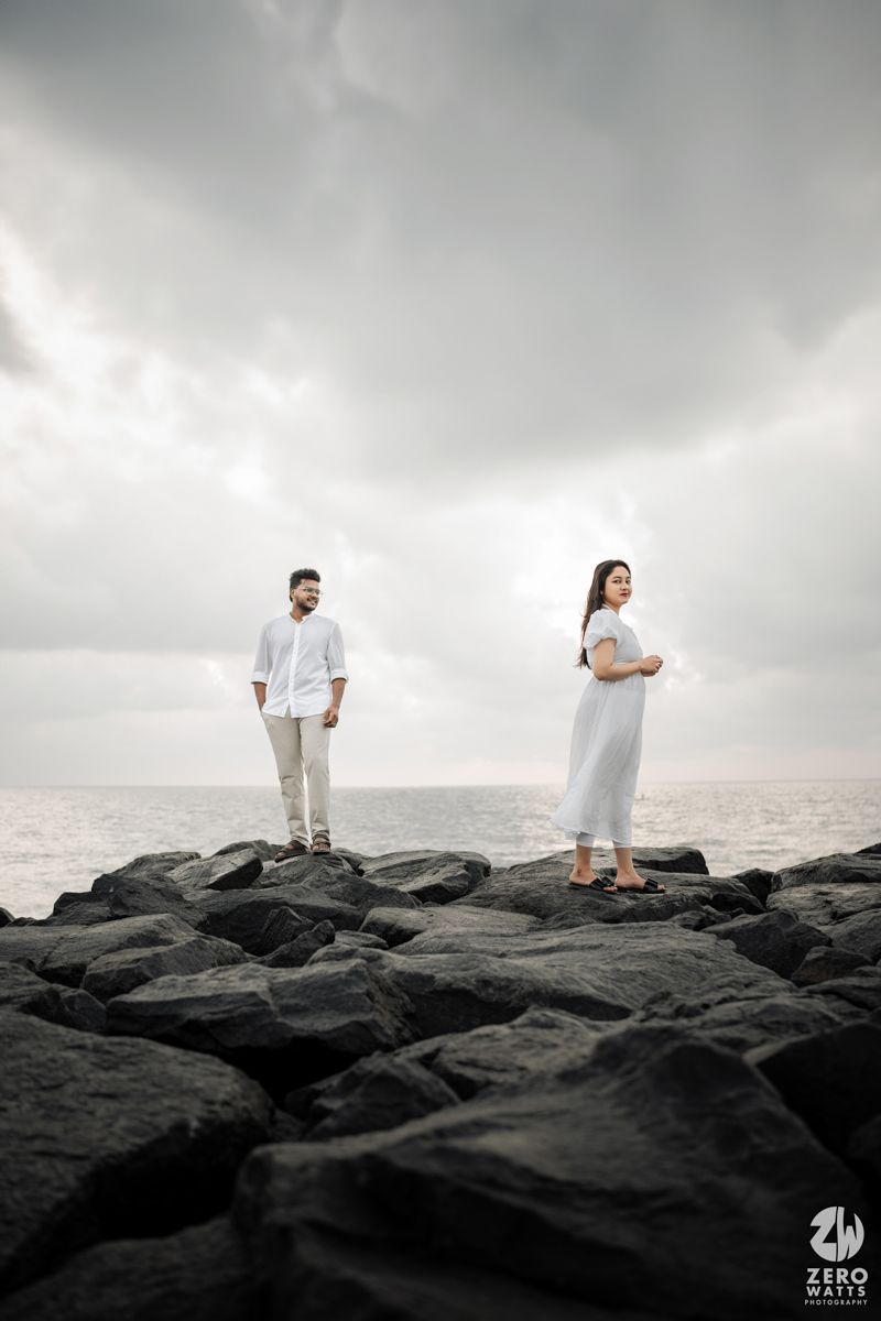 A couple stands apart on dark coastal rocks against cloudy sky and ocean backdrop.