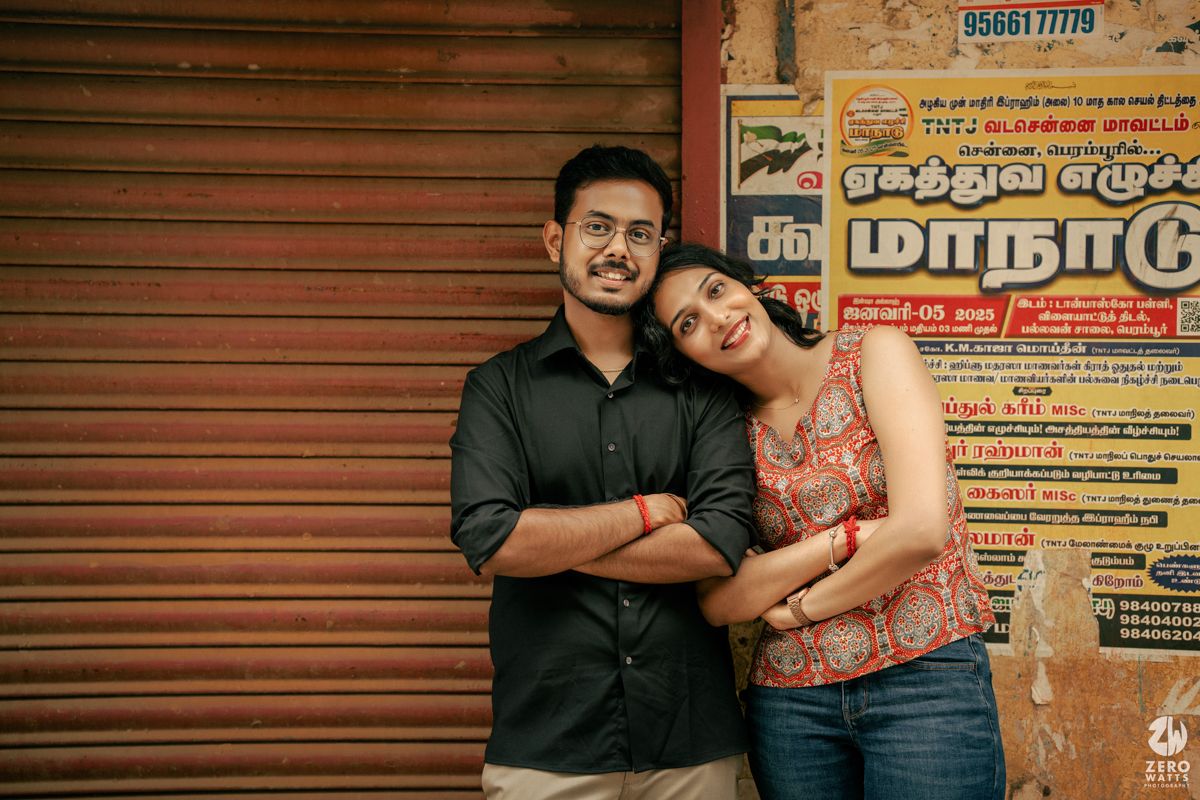 A couple stands casually against a rustic wall with Tamil language posters in the background.