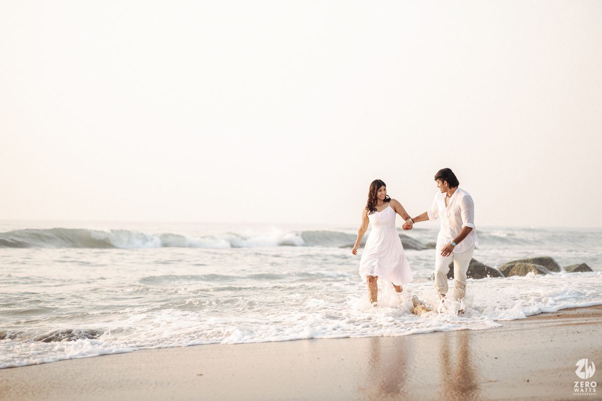 A couple in white outfits walks hand in hand along a beach shore with waves rolling in.