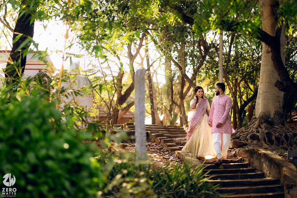 A couple walks down stone steps surrounded by lush green trees and sunlight filtering through the leaves.
