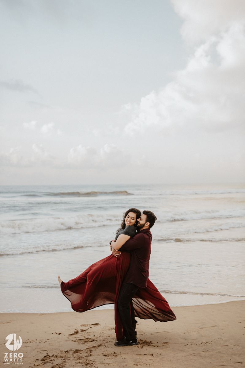 A romantic embrace on the beach with a flowing red dress catching the ocean breeze.