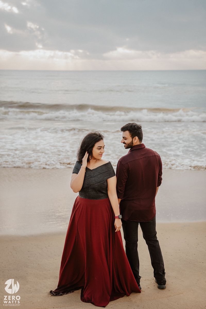 Couple in coordinated burgundy outfits share a tender moment on a cloudy beach.