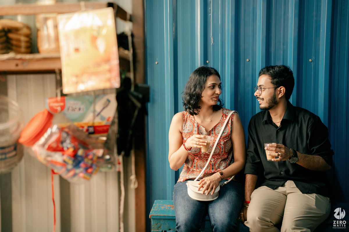 Couple sharing a laugh while leaning against blue corrugated wall with drinks in their hands.