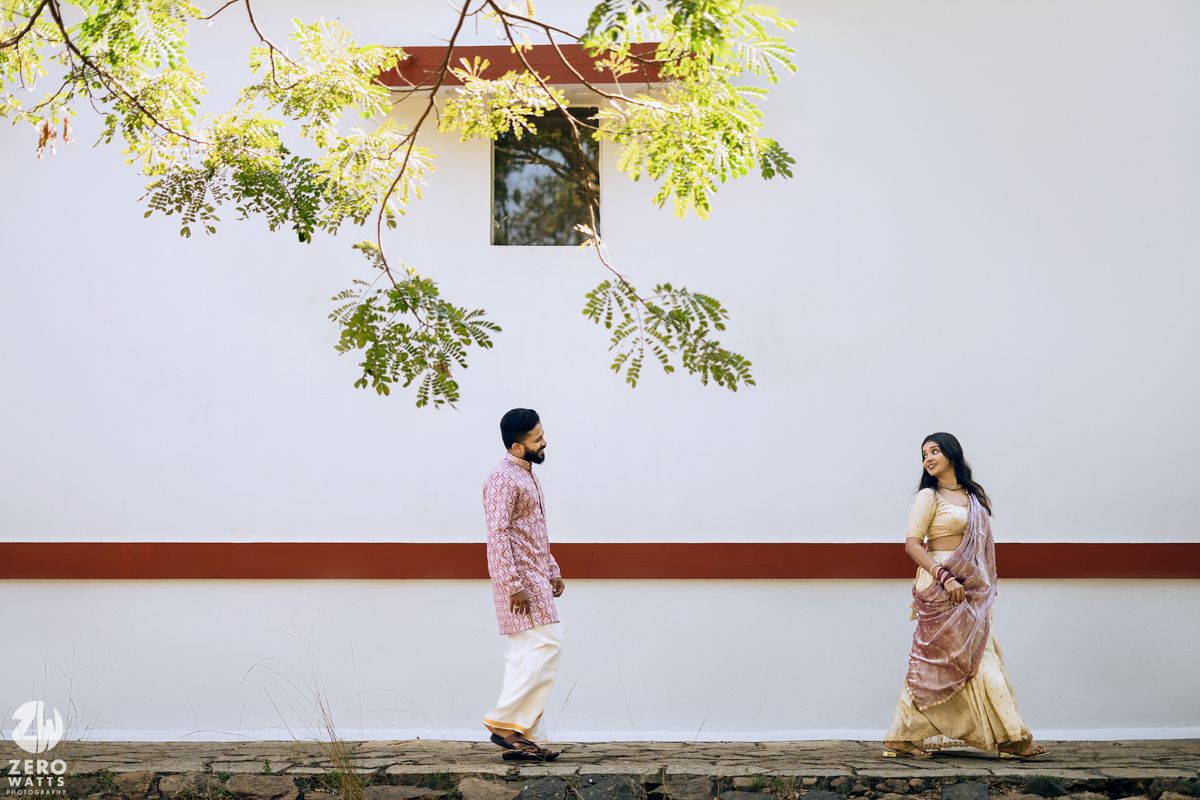 Two people in traditional attire walking along a white wall beneath leafy branches.