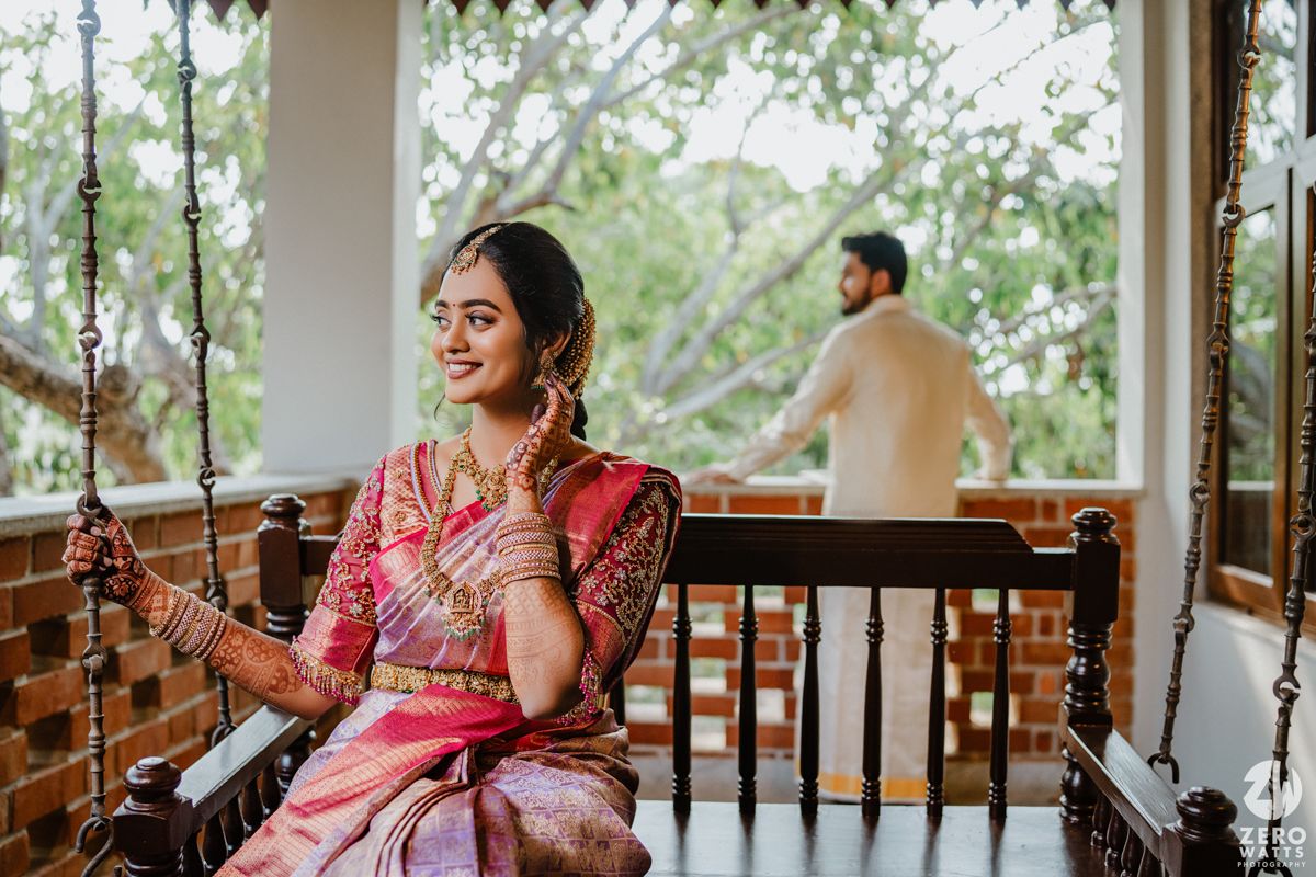 Bride in pink and purple silk saree sits on traditional wooden swing while getting ready for ceremony.